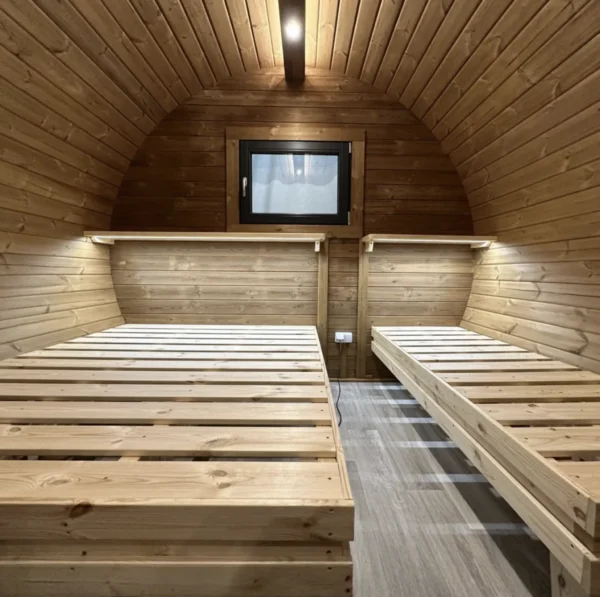 Interior of Camping Barrel 3.0 x 5.9 m featuring wooden walls, two bed frames, cozy lighting, and a small window for ventilation.