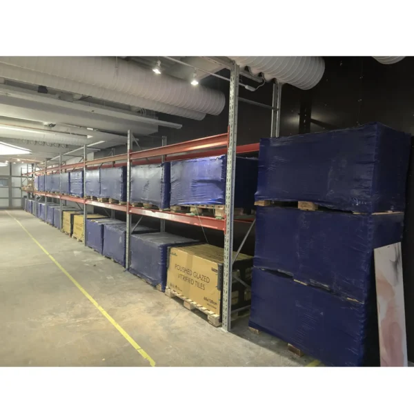 A warehouse interior showing shelves stocked with large packages of porcelain tiles wrapped in blue plastic.