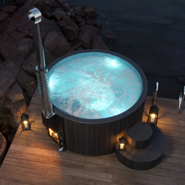 Yoshino Kumano hot tub with dark wooden exterior and blue LED lighting on bubbling water, evening ambiance with lantern lights, silver marine aluminum chimney and wooden deck on rocky shoreline.
